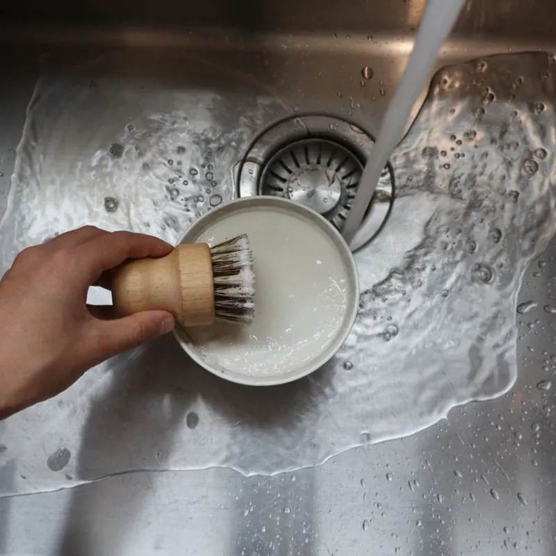 Hand holding brush with dish soap in a sink with running water