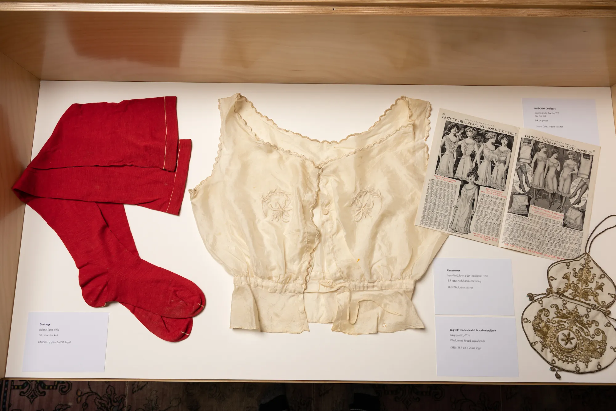 A display case with red stockings, a cream silk corset cover, an open magazine and a decorated bag