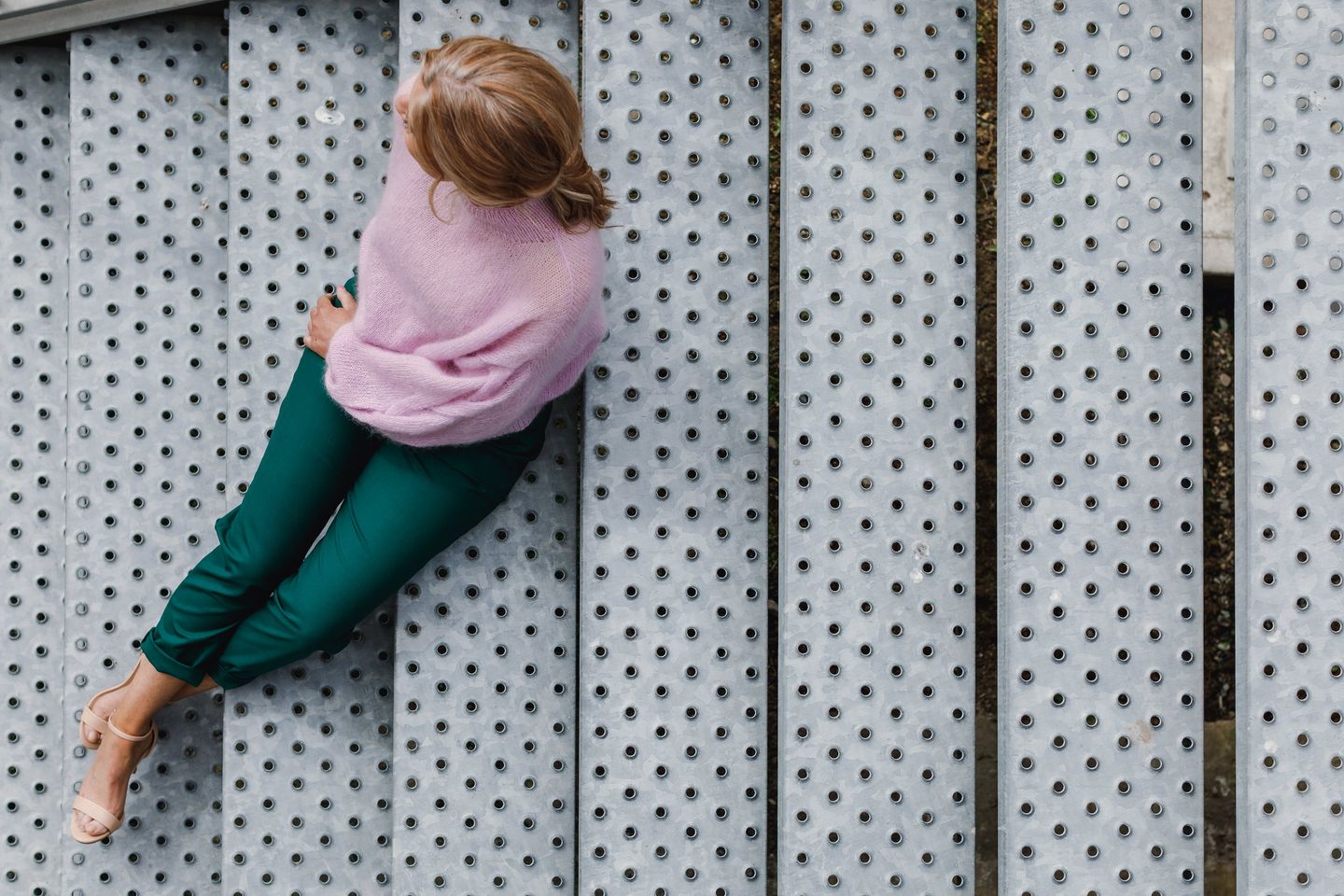 A woman sitting on metal stairs wearing the Plait Sleeve Sweater knitting pattern by Purl Foundry