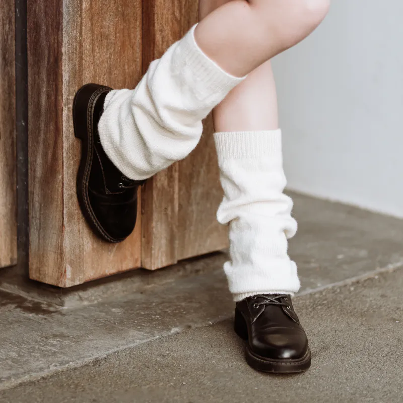 Cream coloured, slouchy leg warmers in plain knitted fabric. The front leg is bent and rests on a wooden post that the model is leaning on.