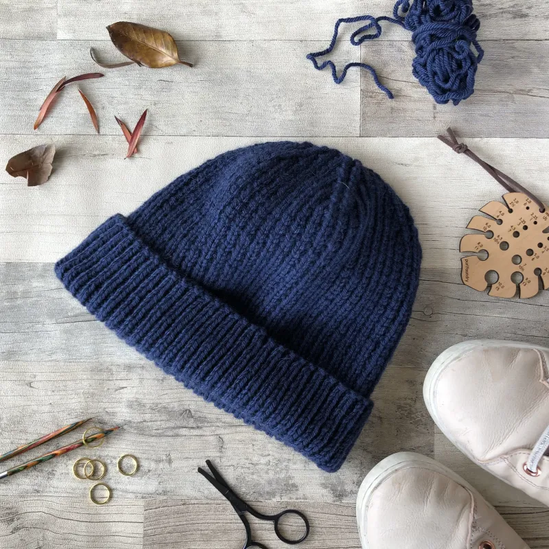 A blue hand knit beanie surrounded by knitting tools