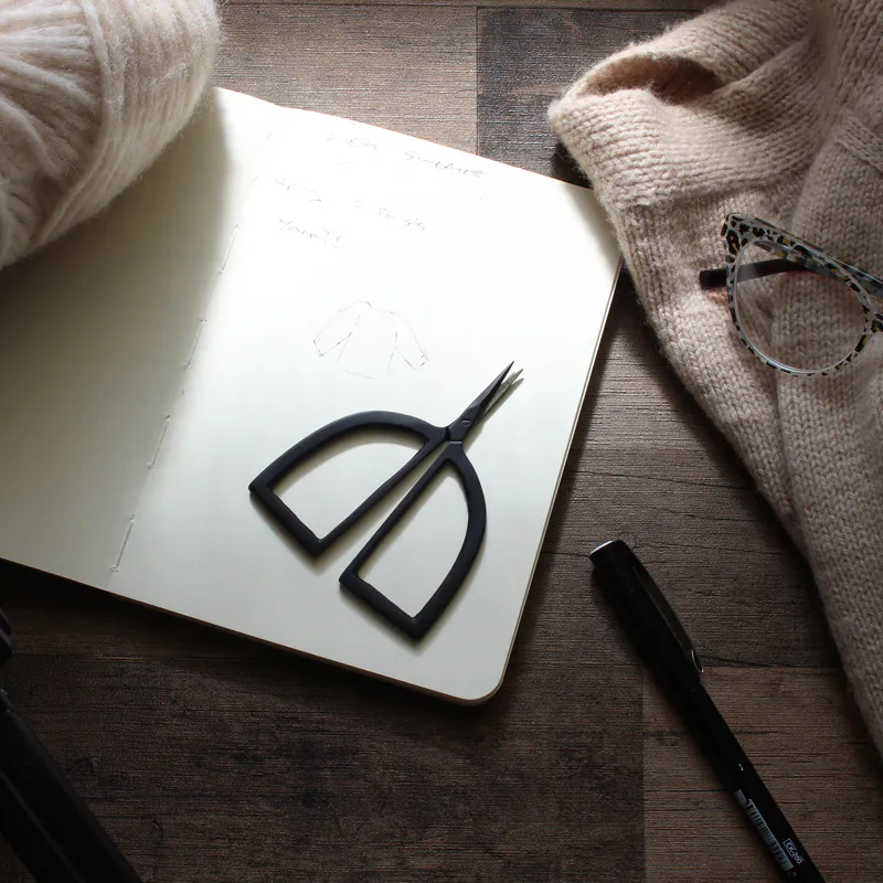 Matte black scissors with arch shaped handles lying on a notebook beside knitting and yarn