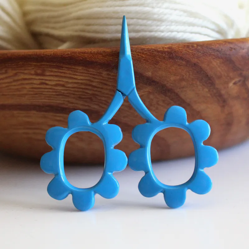 Blue, flower shaped scissors leaning up against a wooden bowl with white coloured wool inside
