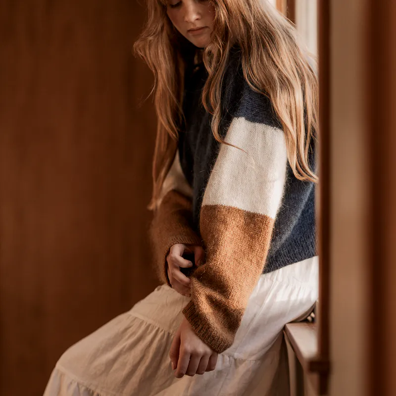 A woman sits on a window ledge and looks down at the cuff on her sweater.