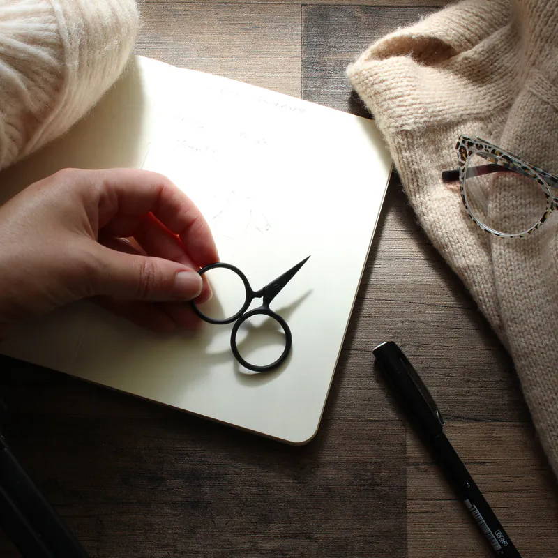 A hand holding black scissors on top of a notebook and surrounded by knitting