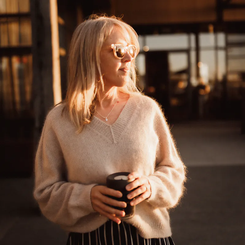 A woman looks to the right into the distance wearing a hand knit, beige sweater called the Ridgeline Sweater. She holds a cup of coffee in front of her.