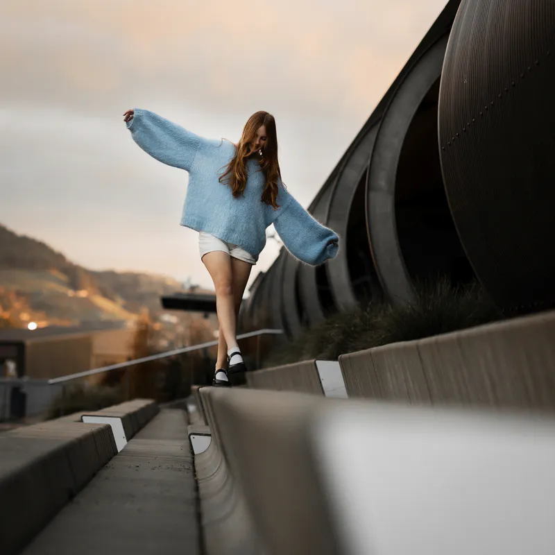 A woman walking along concrete bench seating with her arms spread for balance. She wears a blue mohair jumper