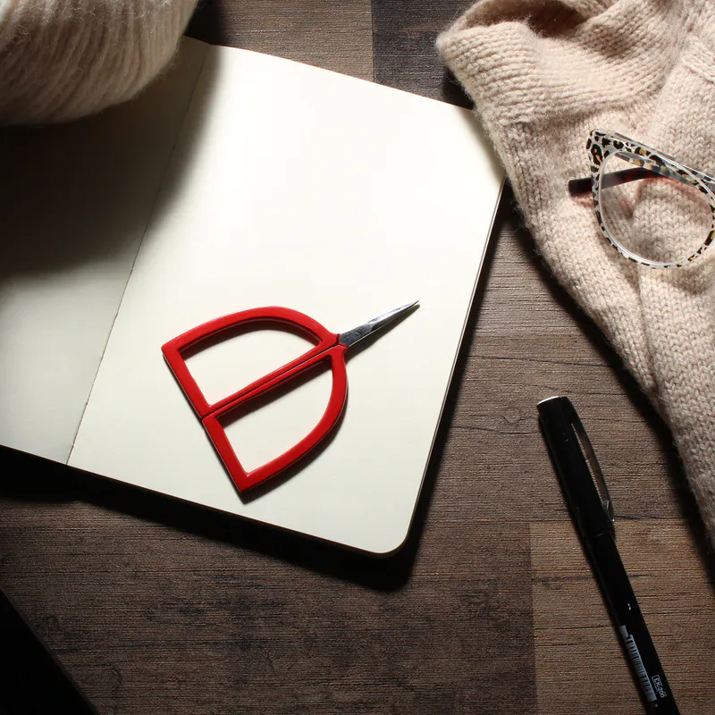 red scissors with arch shaped handles lying on a notebook beside knitting and yarn