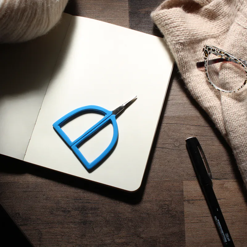 blue scissors with arch shaped handles lying on a notebook beside knitting and yarn