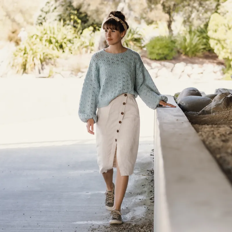 Young woman wearing a blue sweater and linen skirt with sneakers.