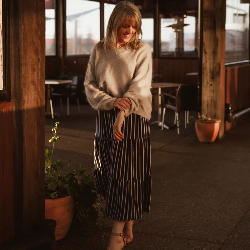 A woman in a beige hand knit sweater gently pushes up the sleeve on one arm. She stands in a cafe area smiling