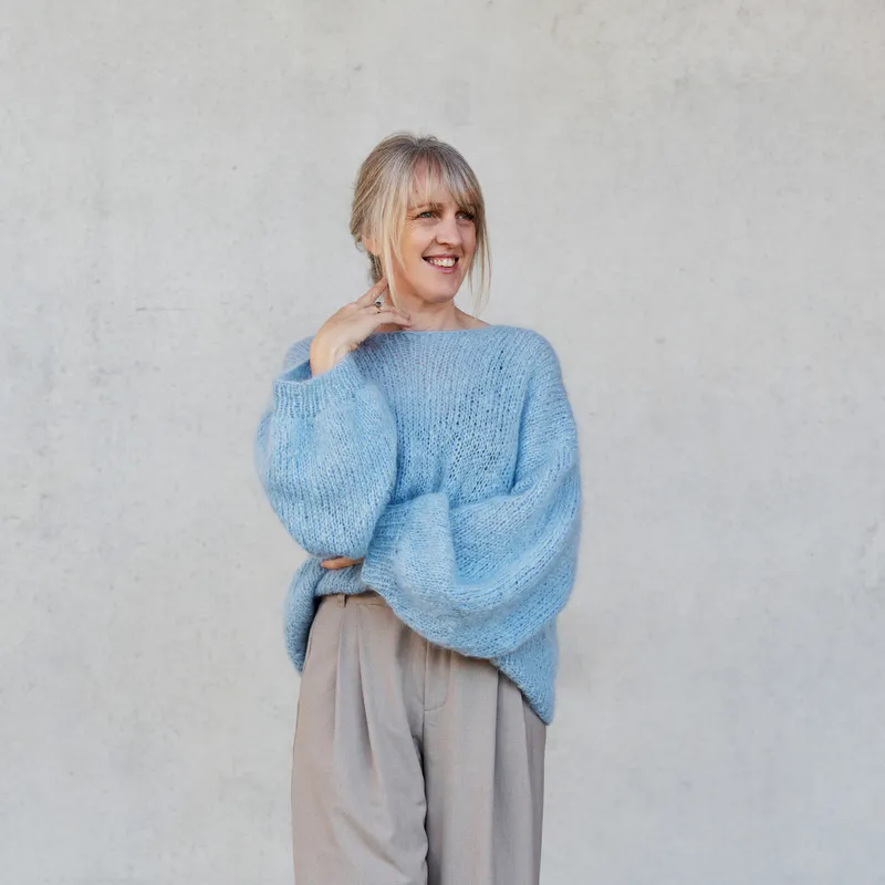 A woman wearing a pale blue mohair sweater which is french tucked into pleated, wide leg pants. Her hand is near her chin with the fingers elegantly splayed