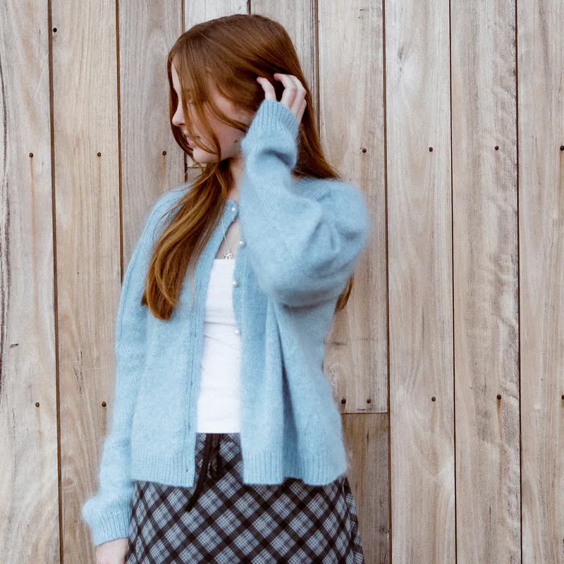 A woman with her hand touching her hair. She wears a cardigan with the top button done up over a white tee and a 90s style skirt.