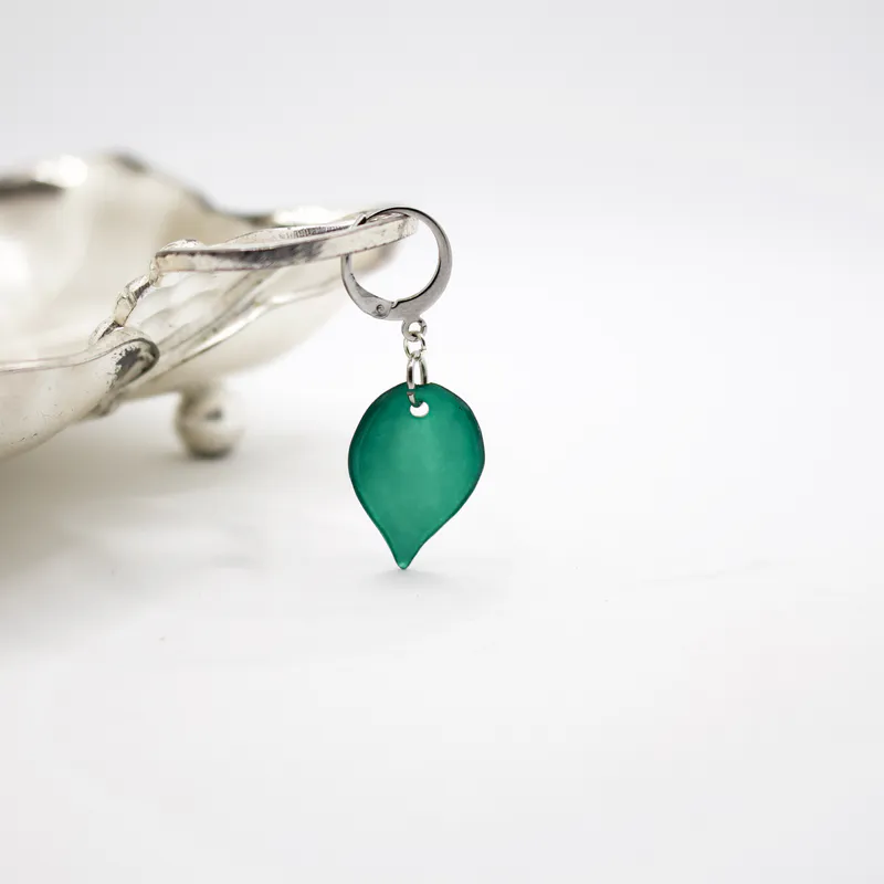 A green acrylic knitting marker in the shape of a leaf. It's hanging from the edge of a thrifted silver dish.
