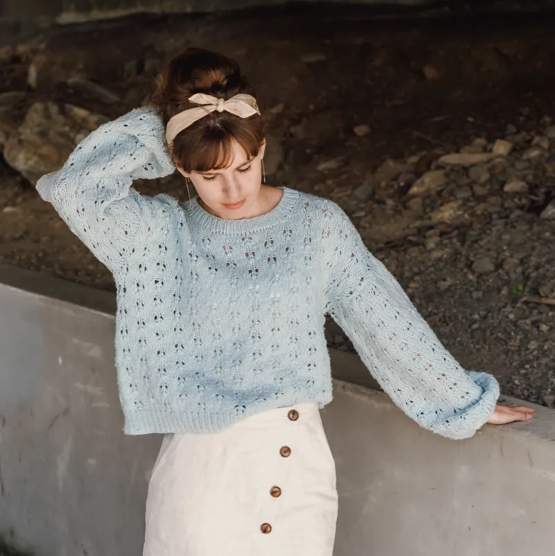 Young woman wearing the Holey Moley Sweater pattern. It's a lace covered, light blue sweater. Her hand is reaching up behind her head and she's looking at the ground.