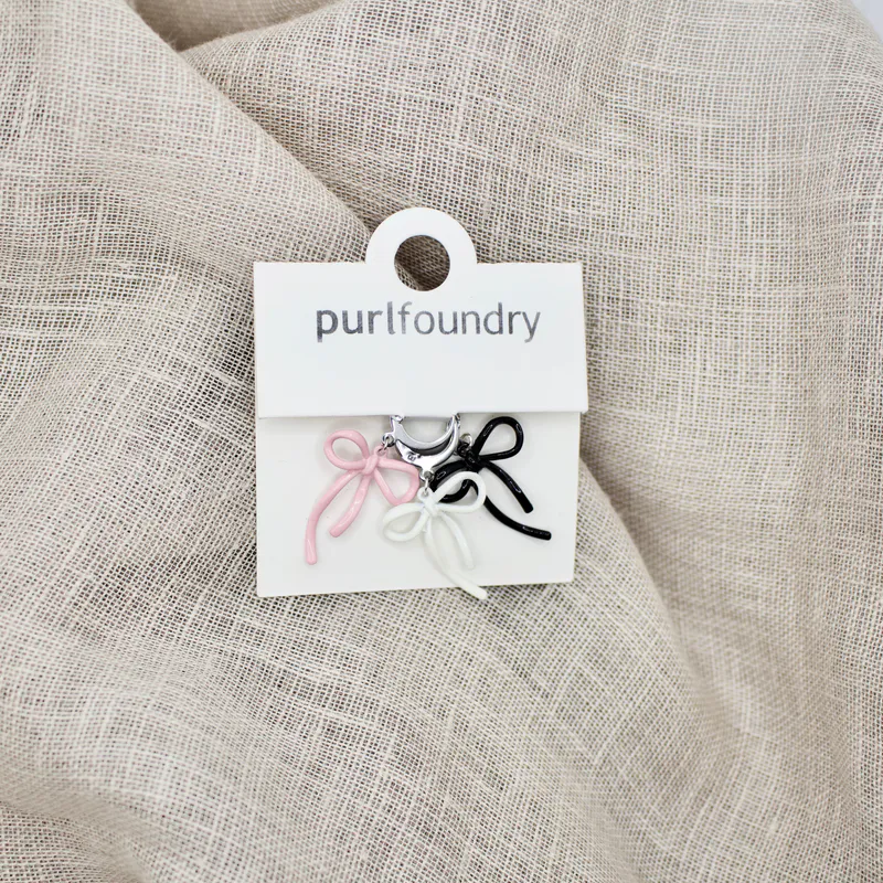 Pink, white and black knitting markers with silver clasps. They come in a pack of three and are packaged on a white card with the Purl Foundry branding. The packaged item sits on some linen.
