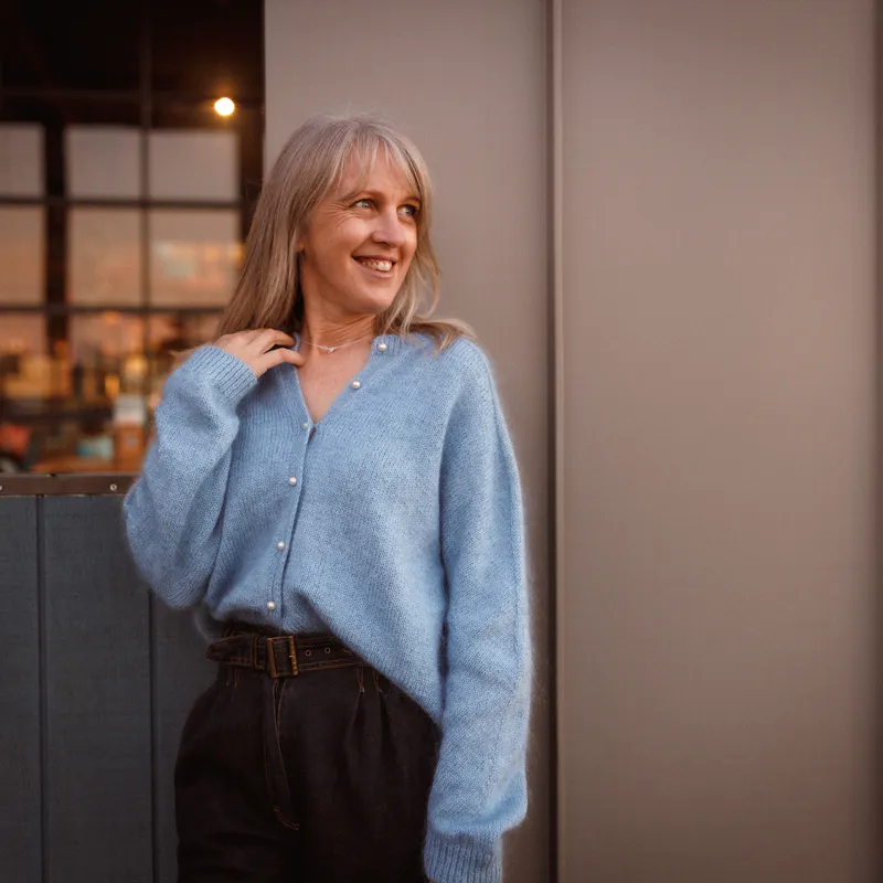 A woman looking to the right with her hand touching her shoulder She wears a pale blue cardigan with pearl buttons and she knit herself which is the best feeling in the world. Plus it's cosy warm!