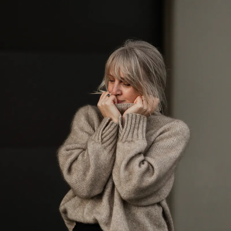 A woman with her hair tied back is holding the edge of a turtleneck to her chin. She smiles softly and looks down. The sweater is an oatmeal colour with a soft fuzz on the fabric