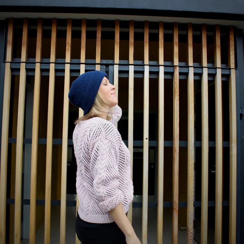 A woman faces upwards, wearing a blue hand knit beanie and a pink hand knit sweater. Her left hand is touching her ear.