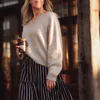 A woman wearing a beige, hand knit sweater. She holds a takeaway coffee in her right hand and a blue and white striped skirt.