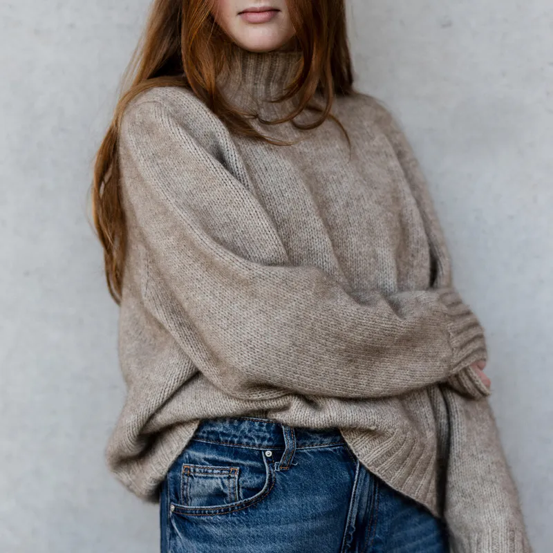 A woman wearing an oversized hand knit in oatmeal coloured yarn. Her arm crosses her body in a one-armed self hug