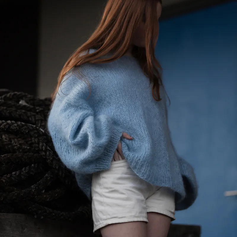 A woman looking away from the camera with a blue door in the background. She wears a pale blue, mohair sweater with cream coloured shorts. One arm is lifting the sweater to place a hand in a pocket