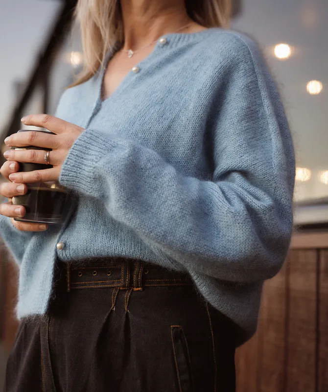 A sky blue, hand knit cardigan with mohair fuzz. It has pearl buttons and a distinct saddle shoulder line running down the length of the sleeve to the cuff