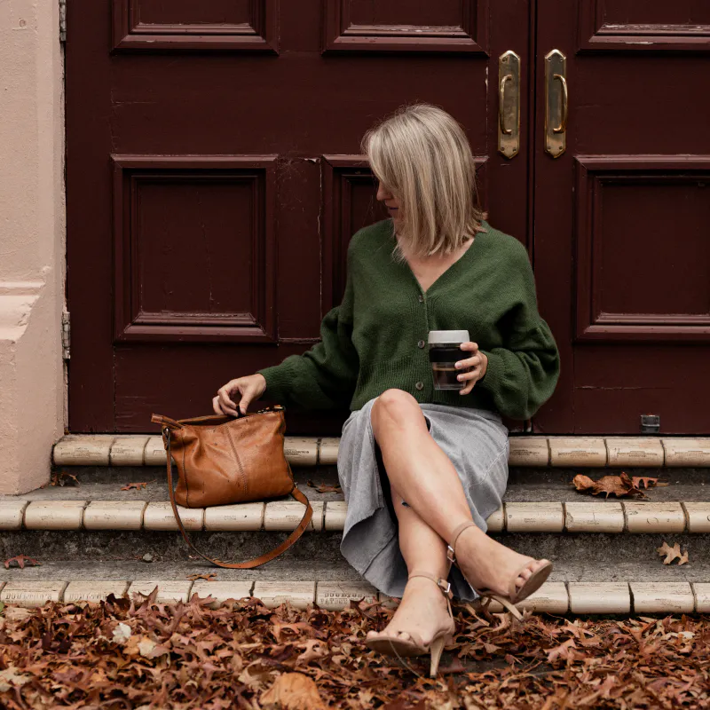 A woman holds coffee in a reusable cup and is rummaging in her brown handbag with other hand. She sits on some steps with her legs crossed and is wearing a green cardigan.