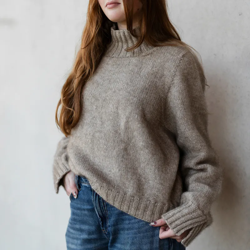 Woman wearing an oatmeal coloured sweater with one hand in her pocket and the other holding the hem. The image is cropped from her chin and you can't see her face.