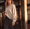 A woman wearing a beige, hand knit sweater. She holds a takeaway coffee in her right hand and a blue and white striped skirt.