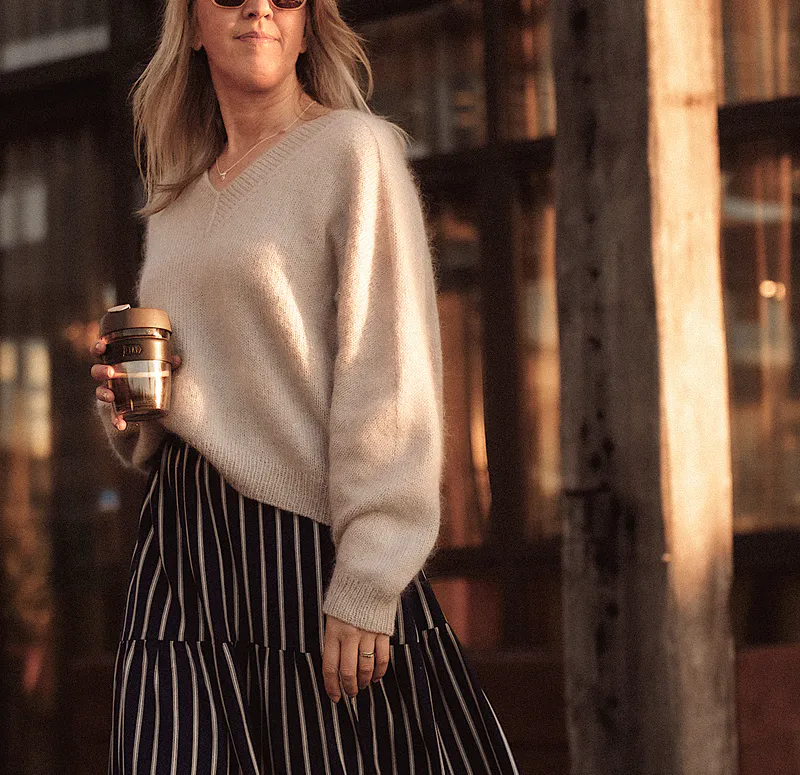 A woman wearing a beige, hand knit sweater. She holds a takeaway coffee in her right hand and a blue and white striped skirt.