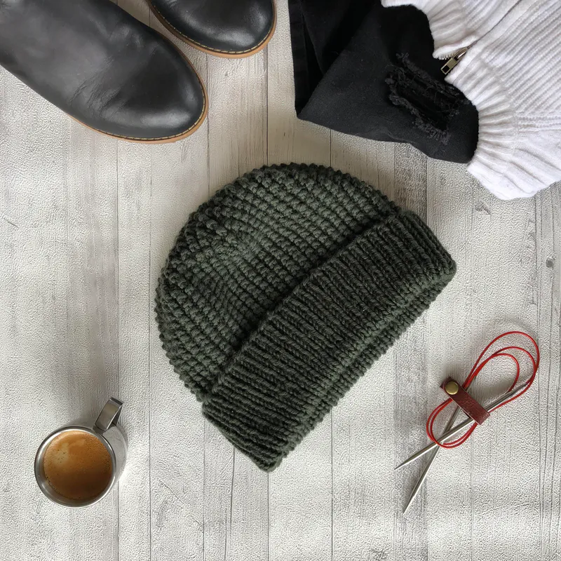 A green knitted hat in the centre with coffee, black boots. clothes and circular knitting needles placed around the edge