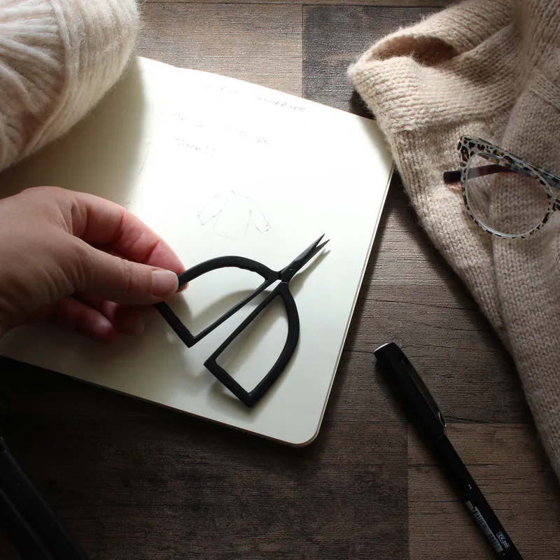 Matte black scissors with arch shaped handles lying on a notebook beside knitting and yarn. A hand is lifting one corner of the scissors