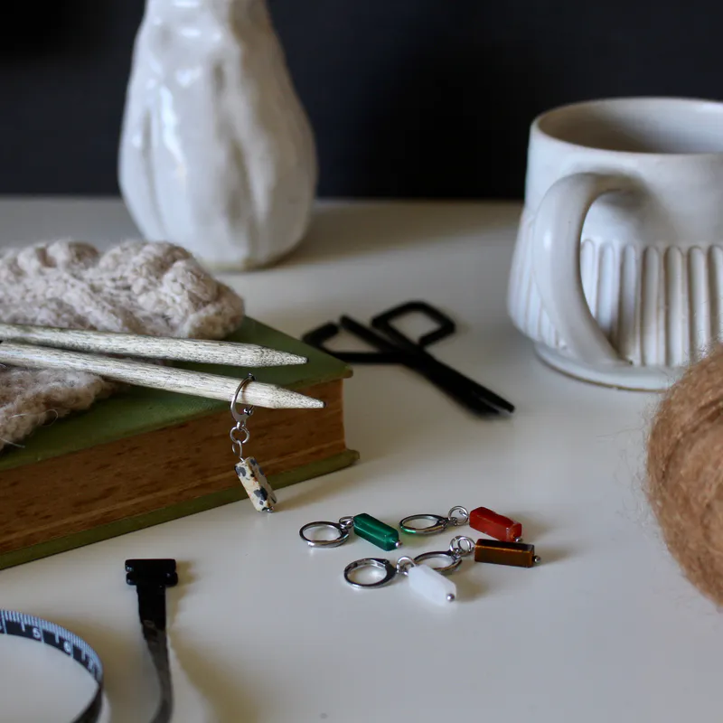 Stone inspired stitch markers on a table. They are surrounded by various items and one stitch marker hangs off a needle which lays on a book
