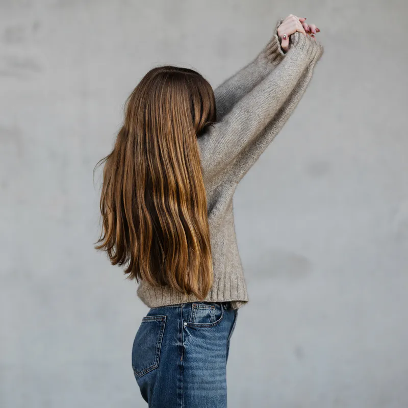 A woman wearing an oatmeal coloured, hand knit sweater with her arms reaching upwards. Her face is covered by her auburn hair.