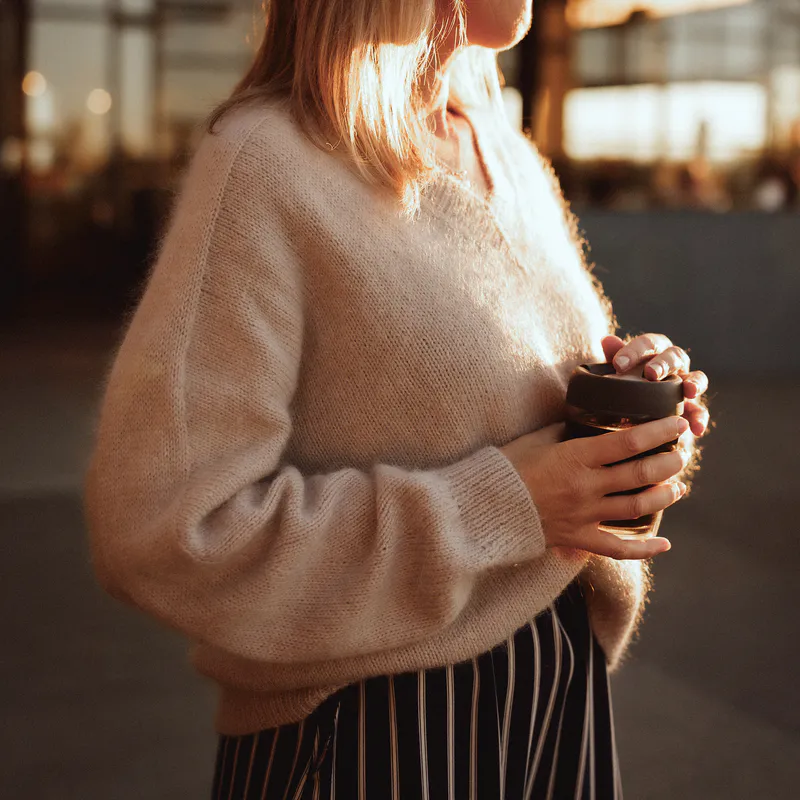A hand knit, beige sweater with the model holding a takeaway coffee in front of her. The takeaway cup is reusable because she loves the environment ... and apparently doing dishes.