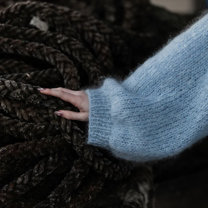 The oversized and draped sleeve and cuff of a hand knit mohair sweater in blue yarn. The hand of the model rests of a messy coil of black rope