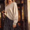 A woman wearing a beige, hand knit sweater. She holds a takeaway coffee in her right hand and a blue and white striped skirt.