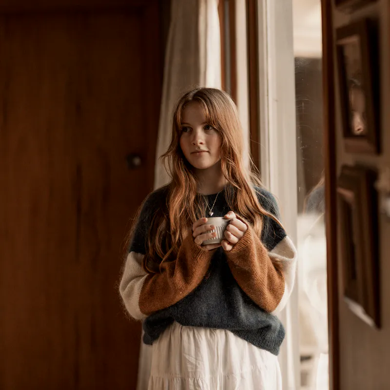 A woman holds a mug close to her chest and looks wistfully to the side. She wears a long white skirt and a hand knit sweater