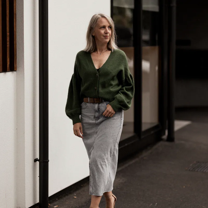 Woman walking down the street wearing a buttoned up V-neck cardigan and a long denim skirt with a slit in the front