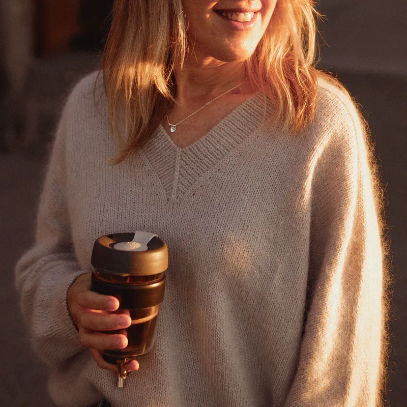 A close up of the v neckline on the Ridgeline Sweater knitting pattern. The beige fabric is fuzzy from the mohair and the woman holds a takeaway coffee in one hand. The sleeve is 100% seamless
