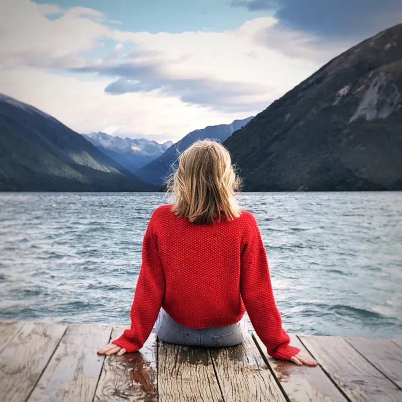 A woman wearing a red sweater with her back to the camera. She sits on a dock overlooking a lake and mountains in New Zealand.