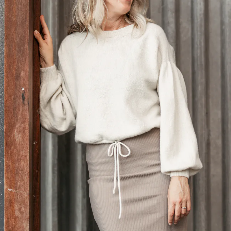 A woman leaning against a wooden column with one arm bent upward the other arm down by her side. She wears an off white, hand knit sweater with a tied waist