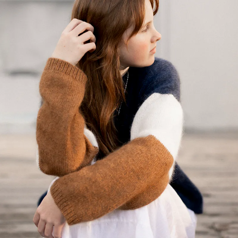 A woman looks away from the camera, wearing a hand knit sweater in three colours. The left arm is playing with her hair.