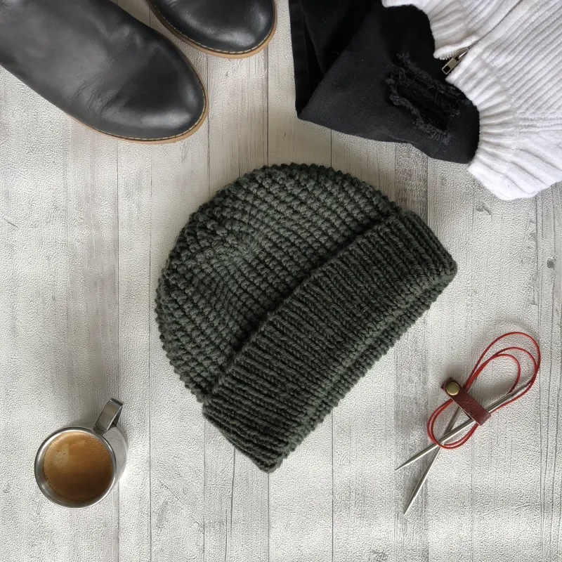A green hand knit beanie laying flat and surrounded by shoes, clothes, knitting needles and a cup of coffee