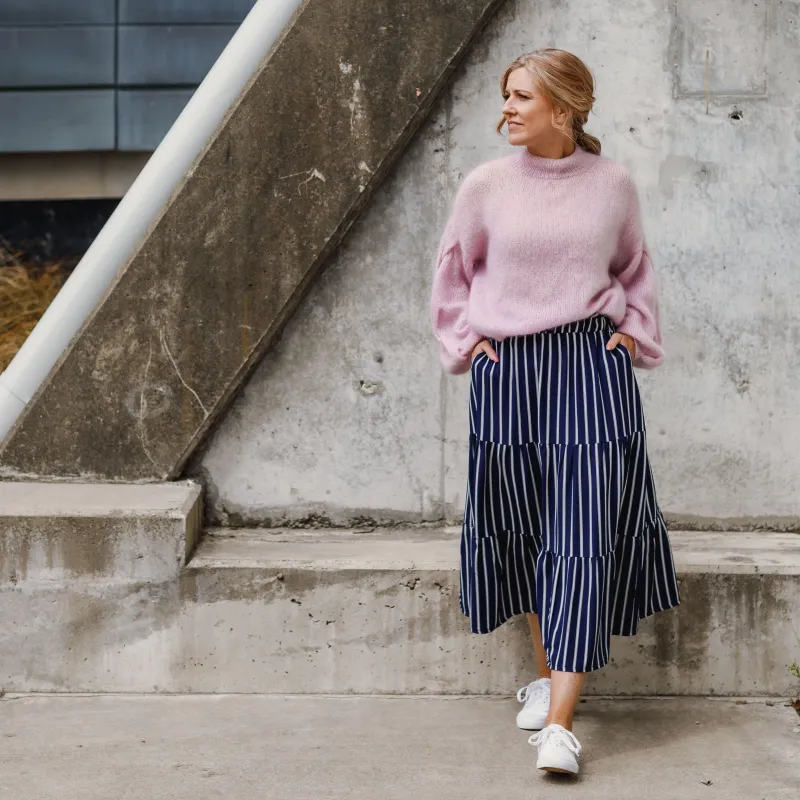 A woman wearing a blue and white striped skirt with tiers, paired with an oversized pink, mohair sweater. The sweater is hand knit.