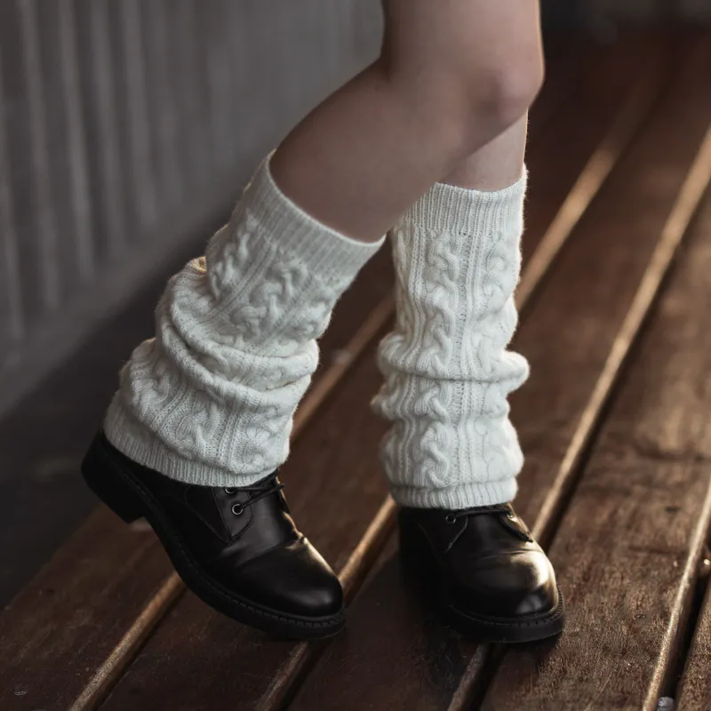 Cream coloured, slouchy leg warmers in cabled knitted fabric. The front leg is bent and the foot is on tip toes.