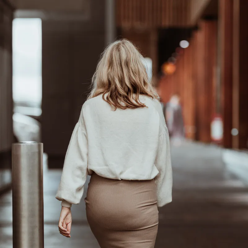 The back view of a hand knit sweater, paired with a dark beige skirt. She has blonde hair and walks down an outdoor, retail alleyway.