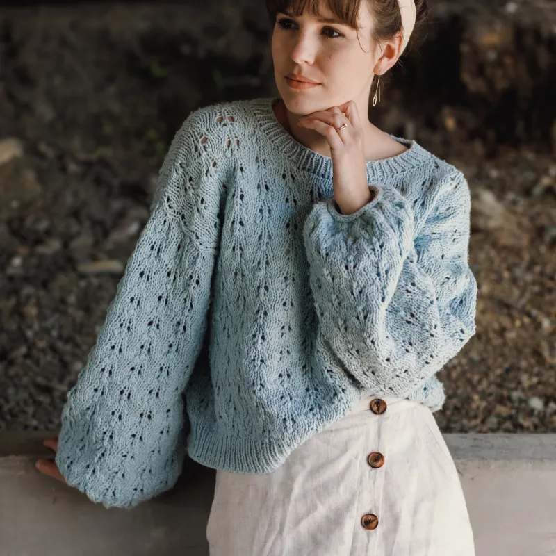 Young woman wearing a blue hand knit sweater with all over lace detail. Her hand is gently positioned under her chin.