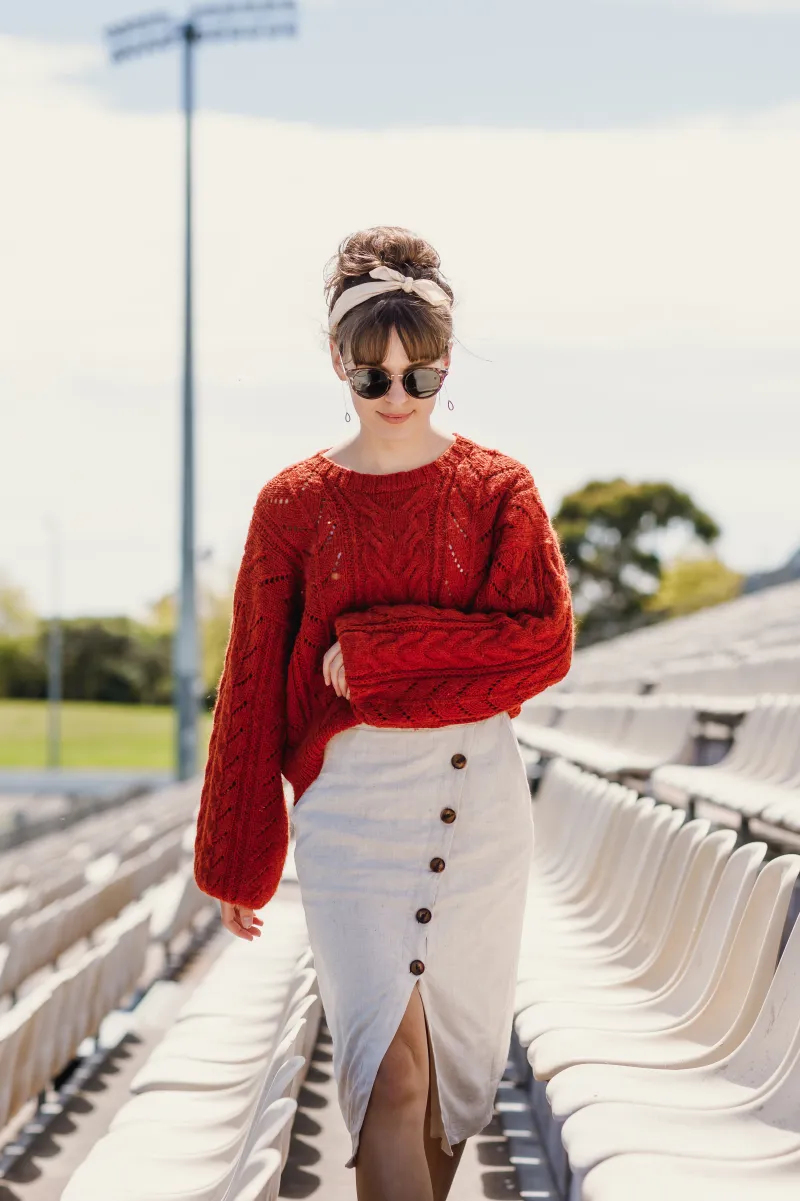 A woman in a linen skirt walks through stadium seating wearing a hand knit sweater with cables. Her arm is crossed in front of her.